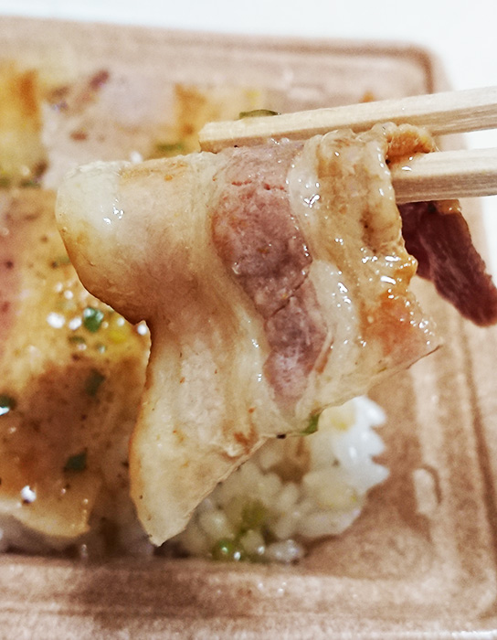 「三元豚のねぎ塩豚カルビ弁当（麦飯）」の豚肉を箸で持ち上げた写真